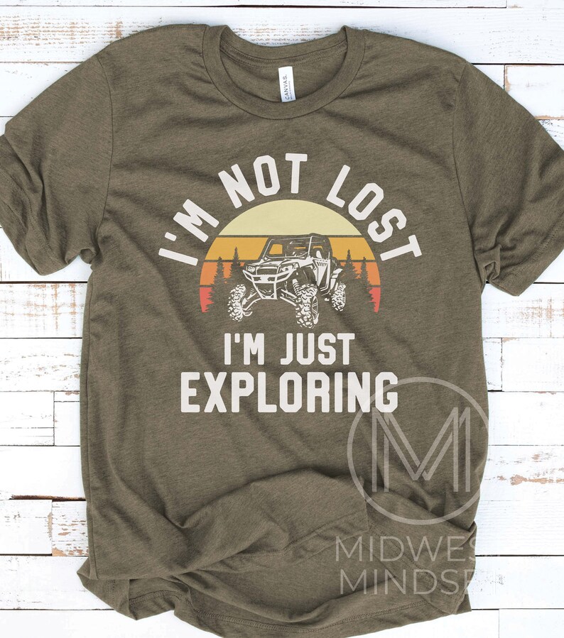 K&ouml;nnte beinhalten: Ein gr&uuml;nes T-Shirt mit einer Retro-Sonnenuntergangsgrafik und einer Silhouette eines Gel&auml;ndewagens. Der Text "Ich bin nicht verloren, ich erkunde nur" ist auf dem Shirt aufgedruckt.