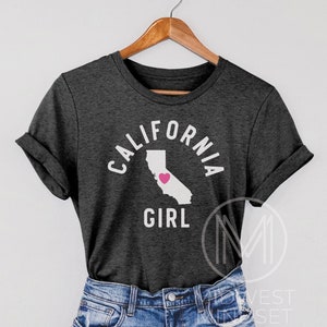 Pu&ograve; includere: Una maglietta grigio scuro con il testo "CALIFORNIA GIRL" in lettere bianche. Il testo &egrave; in un cerchio attorno a un contorno dello stato della California con un cuore rosa al centro.