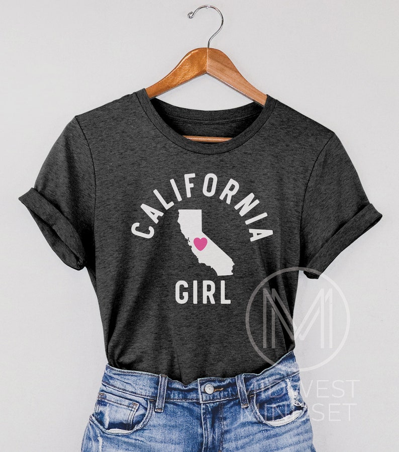 Pu&ograve; includere: Una maglietta grigio scuro con il testo "CALIFORNIA GIRL" in lettere bianche. Il testo &egrave; in un cerchio attorno a un contorno dello stato della California con un cuore rosa al centro.