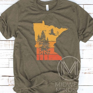 May include: A green t-shirt with a vintage-style graphic of the state of Minnesota. The graphic features a silhouette of the state with a forest and an eagle flying over it. The graphic is in shades of orange, yellow, and red.