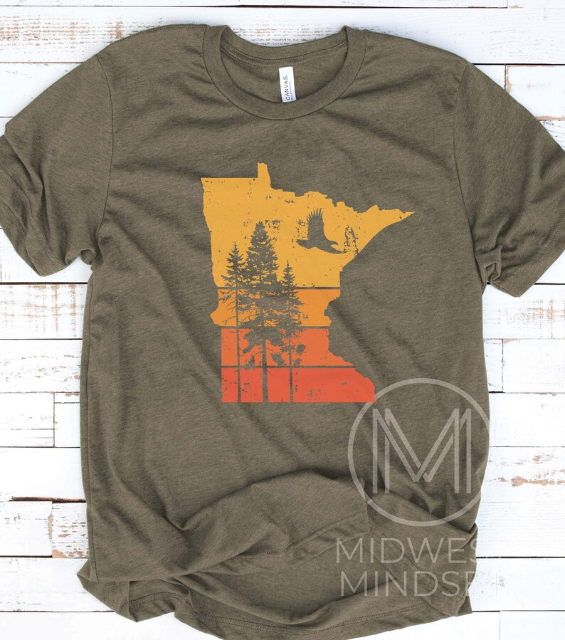 May include: A green t-shirt with a vintage-style graphic of the state of Minnesota. The graphic features a silhouette of the state with a forest and an eagle flying over it. The graphic is in shades of orange, yellow, and red.