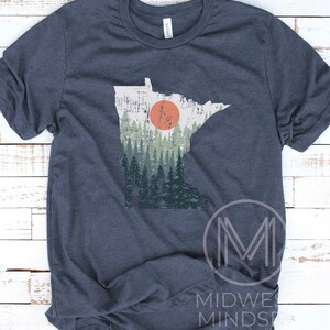 May include: A navy blue t-shirt with a vintage-style graphic of the state of Minnesota. The graphic features a forest scene with a large orange sun in the background.