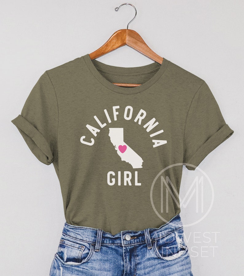 Pu&ograve; includere: Una maglietta verde con il testo "CALIFORNIA GIRL" in lettere bianche. Il testo &egrave; in un cerchio con un contorno dello stato della California all'interno del cerchio. Un cuore rosa &egrave; al centro del contorno dello stato.