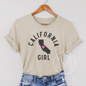 Pu&ograve; includere: Una maglietta beige con il testo "CALIFORNIA GIRL" in lettere nere. Il testo &egrave; in un cerchio attorno a un contorno dello stato della California con un cuore rosa al centro.