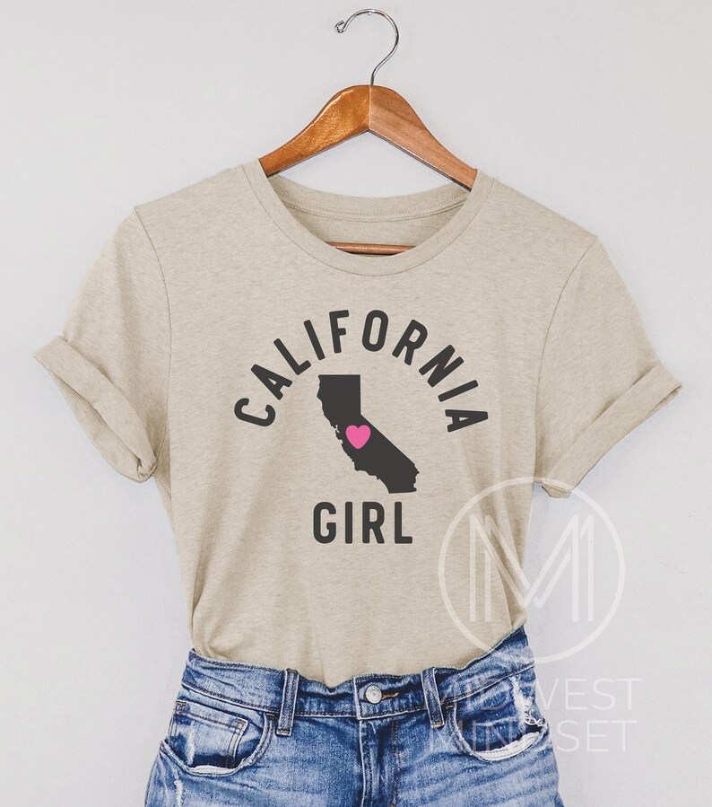 Pu&ograve; includere: Una maglietta beige con il testo "CALIFORNIA GIRL" in lettere nere. Il testo &egrave; in un cerchio attorno a un contorno dello stato della California con un cuore rosa al centro.