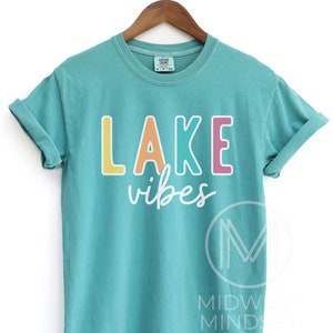 Puede incluir: Camiseta de color turquesa con las palabras "LAKE vibes" en letras de bloque coloridas. La palabra "LAKE" es amarilla, naranja, turquesa y rosa. La palabra "vibes" est&aacute; en una fuente cursiva blanca. La camiseta est&aacute; colgada en una percha de madera.
