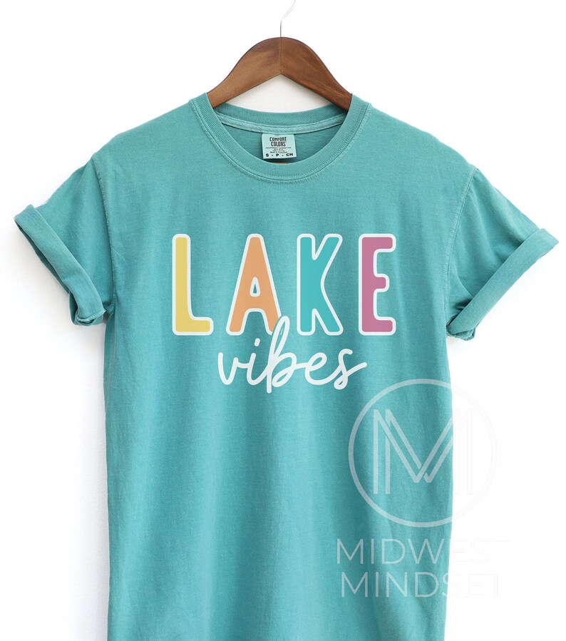 Puede incluir: Camiseta de color turquesa con las palabras "LAKE vibes" en letras de bloque coloridas. La palabra "LAKE" es amarilla, naranja, turquesa y rosa. La palabra "vibes" est&aacute; en una fuente cursiva blanca. La camiseta est&aacute; colgada en una percha de madera.