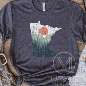 May include: A dark heather grey t-shirt featuring a vintage-style outline of the state of Minnesota with a forest scene and a red sun. The text "MIDWEST MINDS" is printed on the bottom right corner of the shirt.