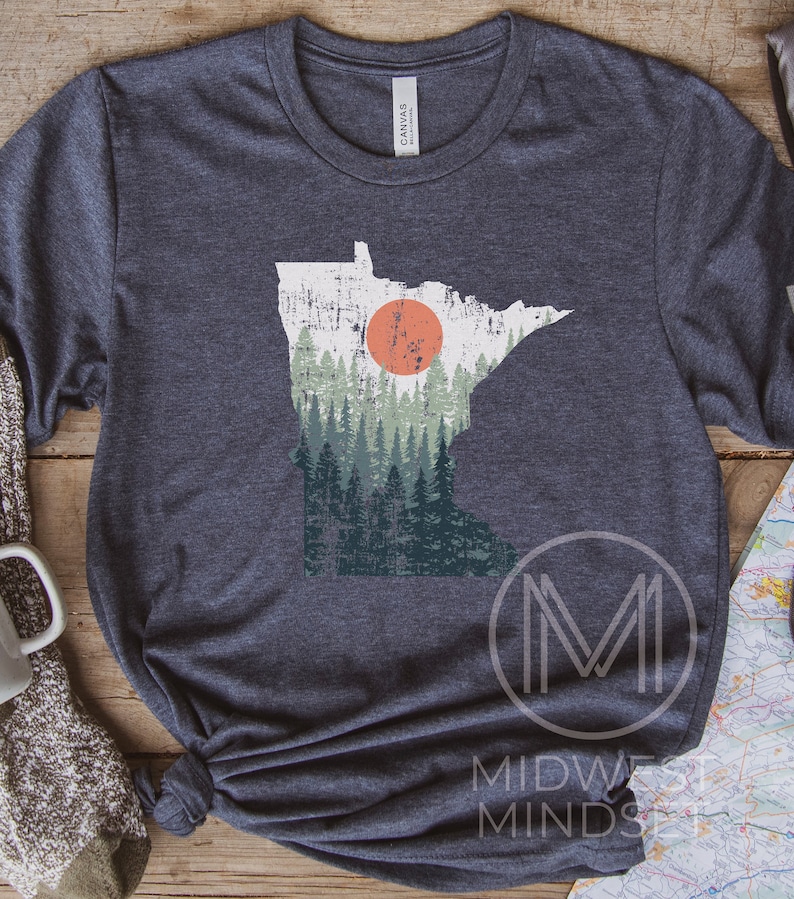 May include: A dark heather grey t-shirt featuring a vintage-style outline of the state of Minnesota with a forest scene and a red sun. The text "MIDWEST MINDS" is printed on the bottom right corner of the shirt.
