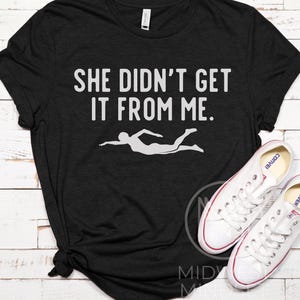 May include: Black t-shirt with white text that reads "She didn't get it from me." and a white silhouette of a person swimming.