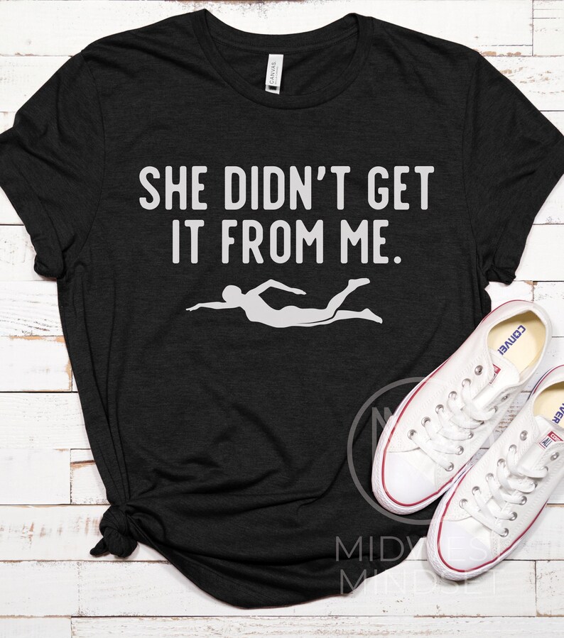May include: Black t-shirt with white text that reads "She didn't get it from me." and a white silhouette of a person swimming.
