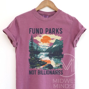 May include: A purple t-shirt with the text "Fund Parks Not Billionaires" and a graphic of a mountain scene with a lake and a sunset.
