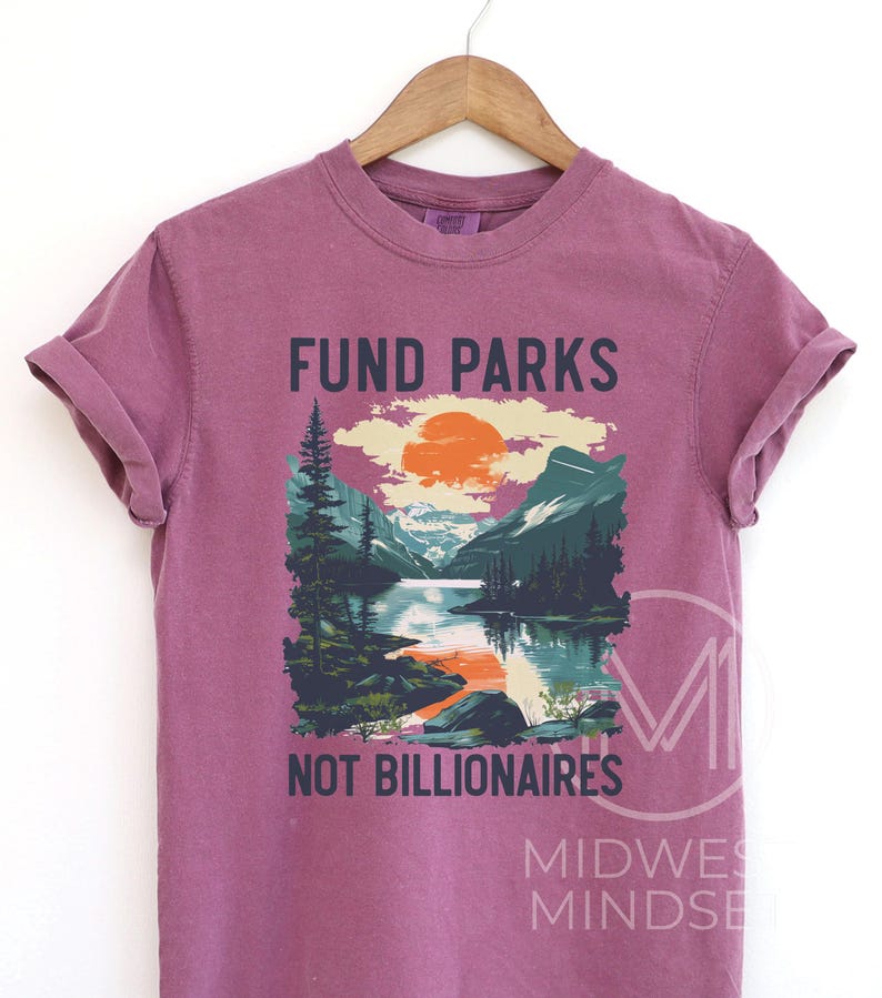 May include: A purple t-shirt with the text "Fund Parks Not Billionaires" and a graphic of a mountain scene with a lake and a sunset.