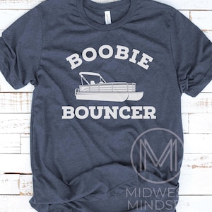 May include: A navy blue t-shirt with white text that reads "Boobie Bouncer" and a white silhouette of a pontoon boat. The shirt is made of a soft, comfortable material.
