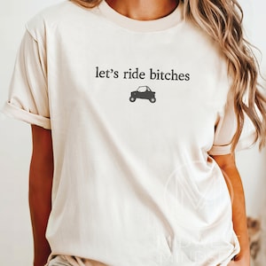 Puede incluir: Camiseta color crema con la frase "let's ride bitches" en letras grises oscuras sobre un peque&ntilde;o gr&aacute;fico negro de un veh&iacute;culo todoterreno. La camiseta tiene mangas cortas y un corte holgado.