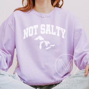 May include: A lavender sweatshirt with the text "NOT SALTY" in white, featuring a white outline of the Great Lakes. The sweatshirt is worn by a person sitting with their legs crossed.