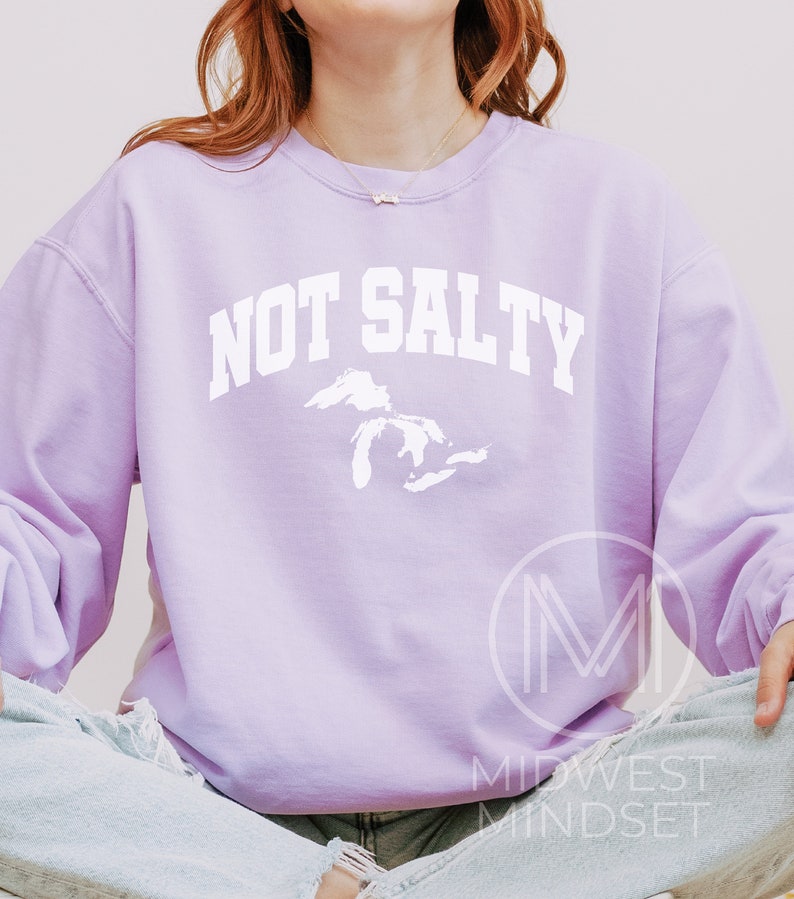 May include: A lavender sweatshirt with the text "NOT SALTY" in white, featuring a white outline of the Great Lakes. The sweatshirt is worn by a person sitting with their legs crossed.