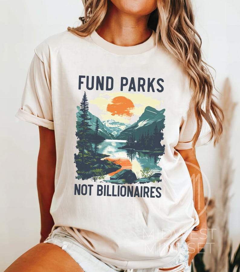 May include: A beige t-shirt with a graphic design of a mountain landscape with a setting sun. The text "FUND PARKS NOT BILLIONAIRES" is printed on the shirt.