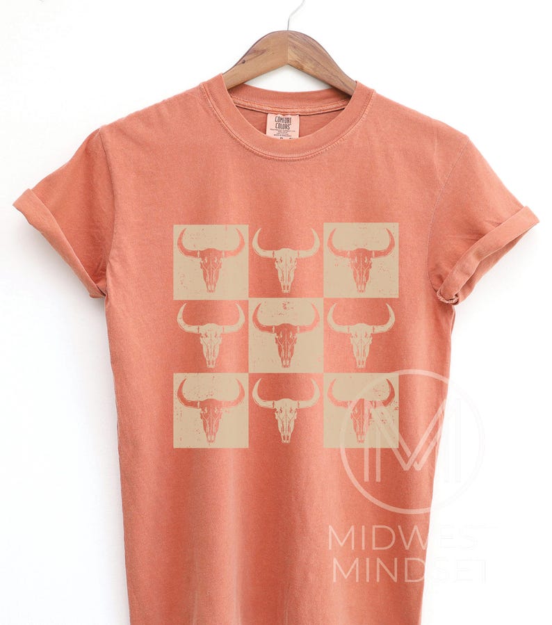 Pu&ograve; includere: Una maglietta color corallo arancio con un motivo di nove teschi di toro bianchi. I teschi sono disposti in una griglia 3x3.