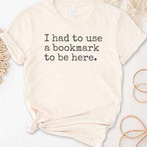 May include: Cream-colored t-shirt with the text "I had to use a bookmark to be here." The shirt has a crew neck and short sleeves, with a knot at the bottom. The shirt is made of a soft material.