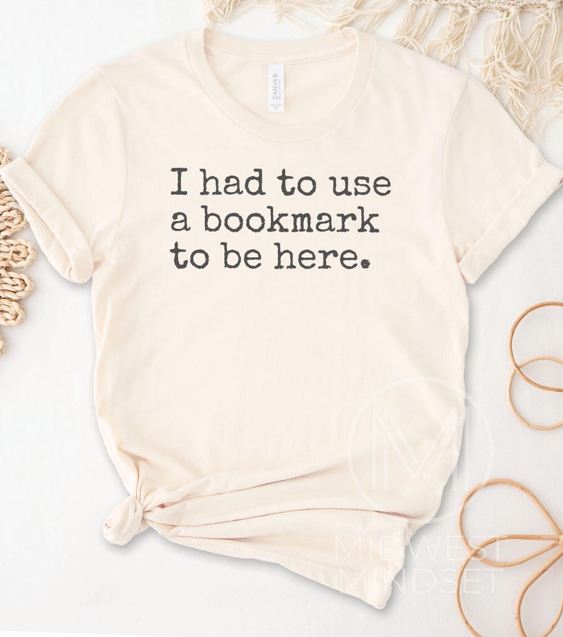 May include: Cream-colored t-shirt with the text "I had to use a bookmark to be here." The shirt has a crew neck and short sleeves, with a knot at the bottom. The shirt is made of a soft material.