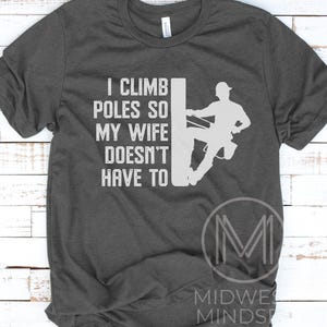 Puede incluir: Una camiseta gris oscuro con un gr&aacute;fico blanco que dice "Subo a los postes para que mi esposa no tenga que hacerlo". El gr&aacute;fico tambi&eacute;n incluye una silueta de una persona subiendo a un poste.