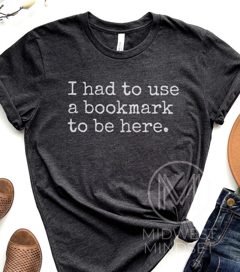 May include: Dark gray t-shirt with the text "I had to use a bookmark to be here" in white. The shirt is a crew neck style and appears to be made of a soft material. The shirt is slightly wrinkled.