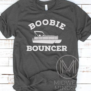 May include: A dark gray t-shirt with the text "Boobie Bouncer" in white. A white silhouette of a pontoon boat is below the text.