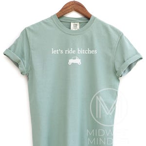 Puede incluir: Camiseta verde claro con la frase blanca "let's ride bitches" y un peque&ntilde;o gr&aacute;fico de coche. La camiseta est&aacute; colgada en una percha de madera.