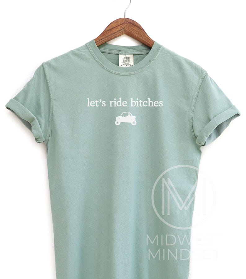 Puede incluir: Camiseta verde claro con la frase blanca "let's ride bitches" y un peque&ntilde;o gr&aacute;fico de coche. La camiseta est&aacute; colgada en una percha de madera.