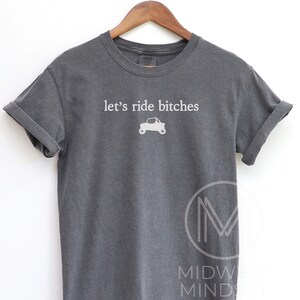 Puede incluir: Camiseta gris oscuro con mangas enrolladas. La camiseta presenta la frase "let's ride bitches" en blanco, junto con un peque&ntilde;o gr&aacute;fico de un coche. Cuelga de una percha de madera.