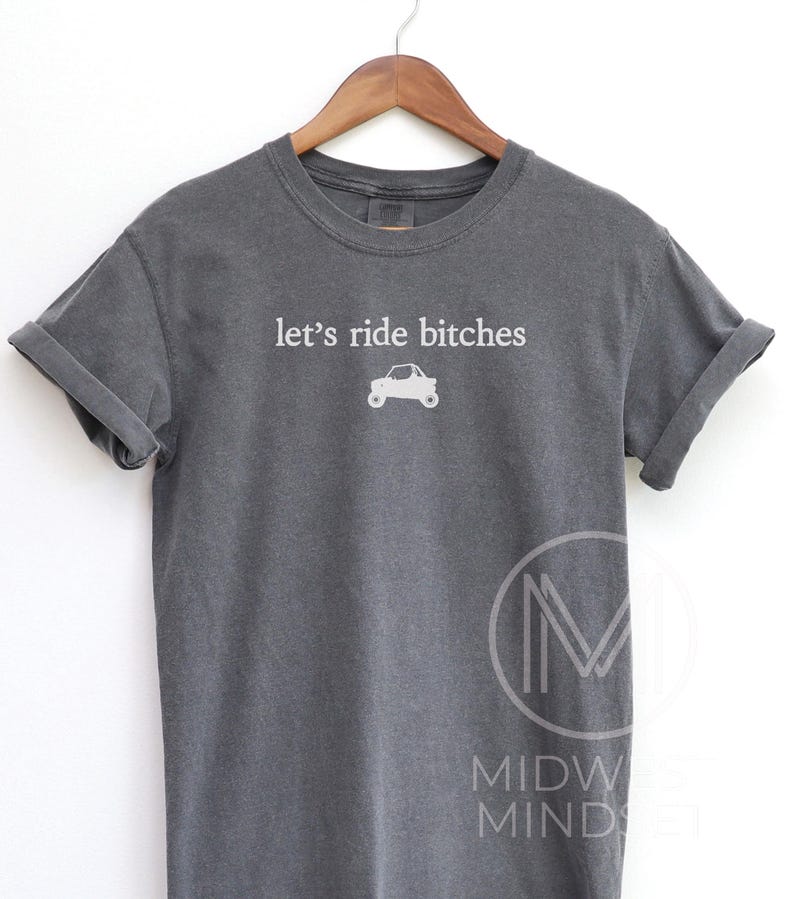 Puede incluir: Camiseta gris oscuro con mangas enrolladas. La camiseta presenta la frase "let's ride bitches" en blanco, junto con un peque&ntilde;o gr&aacute;fico de un coche. Cuelga de una percha de madera.