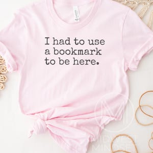 May include: A light pink t-shirt with the text "I had to use a bookmark to be here" in black. The shirt is knotted at the bottom and has a crew neck. The shirt is made of a soft material.