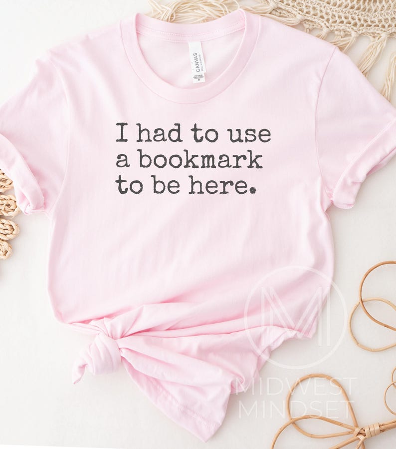 May include: A light pink t-shirt with the text "I had to use a bookmark to be here" in black. The shirt is knotted at the bottom and has a crew neck. The shirt is made of a soft material.