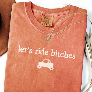 May include: Coral-colored t-shirt with the phrase "let's ride bitches" and a graphic of an off-road vehicle. A gold necklace with a pendant rests on the shirt. The shirt has a Comfort Colors tag.