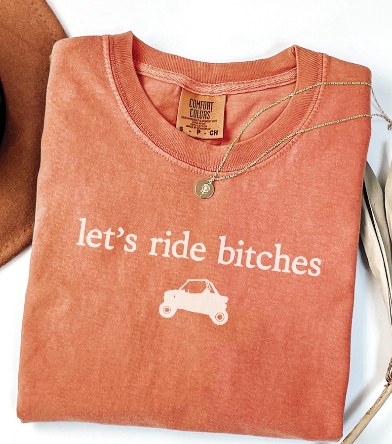 Puede incluir: Camiseta de color coral con la frase "let's ride bitches" y un gr&aacute;fico de un veh&iacute;culo todoterreno. Un collar de oro con un colgante descansa sobre la camiseta. La camiseta tiene una etiqueta Comfort Colors.