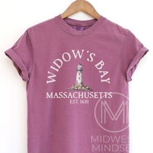 May include: A mauve short-sleeve t-shirt featuring a graphic of a lighthouse on rocks with the text WIDOW'S BAY, MASSACHUSETTS, and EST. 1681 printed in white. The shirt has a crew neckline and is displayed on a wooden hanger against a plain background.