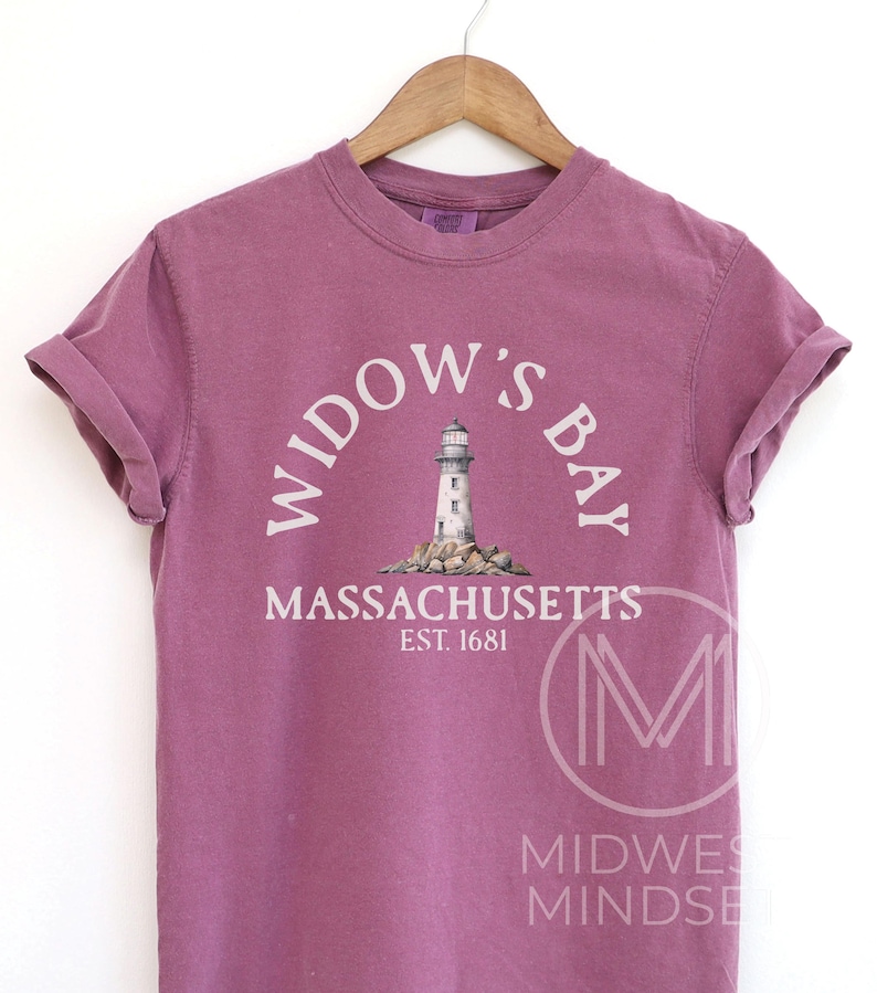 May include: A mauve short-sleeve t-shirt featuring a graphic of a lighthouse on rocks with the text WIDOW'S BAY, MASSACHUSETTS, and EST. 1681 printed in white. The shirt has a crew neckline and is displayed on a wooden hanger against a plain background.