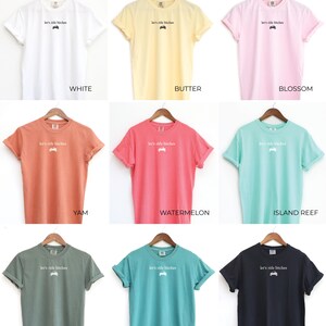 Puede incluir: Una colecci&oacute;n de nueve camisetas de manga corta en varios colores, incluyendo blanco, amarillo, rosa, naranja, rojo, verde azulado, verde, azul y negro. Cada camiseta tiene el texto "let's ride bitches" y un peque&ntilde;o gr&aacute;fico.