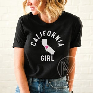 Pu&ograve; includere: Maglietta nera con testo bianco che recita "CALIFORNIA GIRL" e un contorno bianco dello stato della California con un cuore rosa al centro.