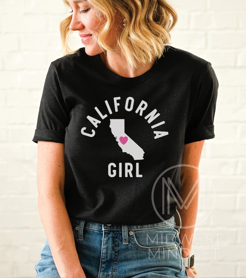 Pu&ograve; includere: Maglietta nera con testo bianco che recita "CALIFORNIA GIRL" e un contorno bianco dello stato della California con un cuore rosa al centro.