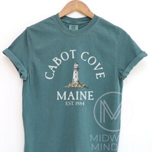 以下が含まれることがあります： 「Cabot Cove Maine」の文字がアーチ状に配置され、灯台のグラフィックが描かれた、ティール色のTシャツ。「MAINE EST. 1984」の文字が下にあります。木製のハンガーに掛けられています。