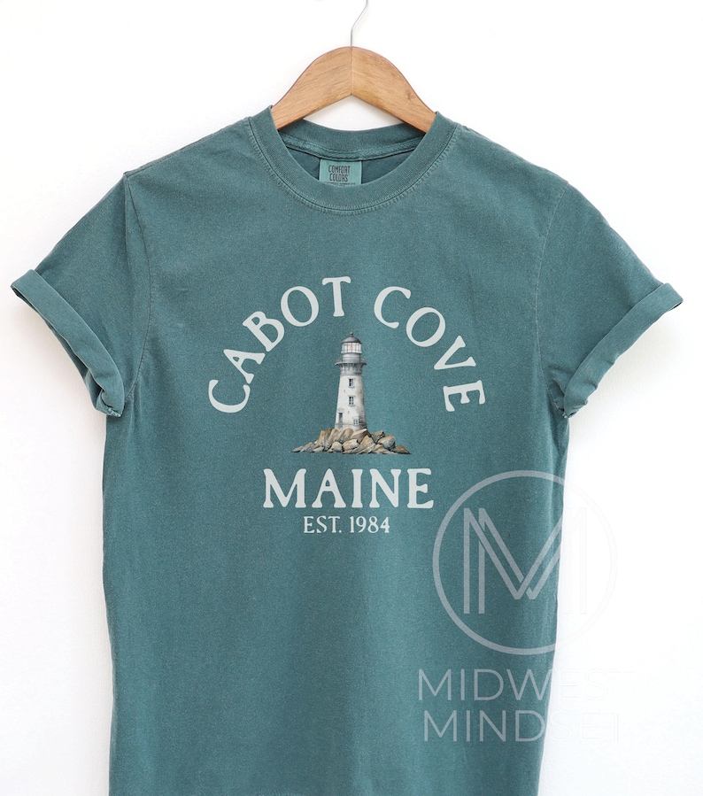 以下が含まれることがあります： 「Cabot Cove Maine」の文字がアーチ状に配置され、灯台のグラフィックが描かれた、ティール色のTシャツ。「MAINE EST. 1984」の文字が下にあります。木製のハンガーに掛けられています。