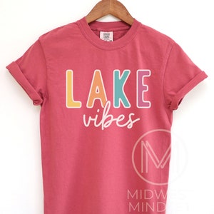 Puede incluir: Una camiseta roja de manga corta con las palabras "LAKE vibes" impresas en la parte delantera en letras coloridas. La camiseta est&aacute; colgada en una percha de madera sobre un fondo blanco. El texto est&aacute; en una fuente l&uacute;dica e informal.