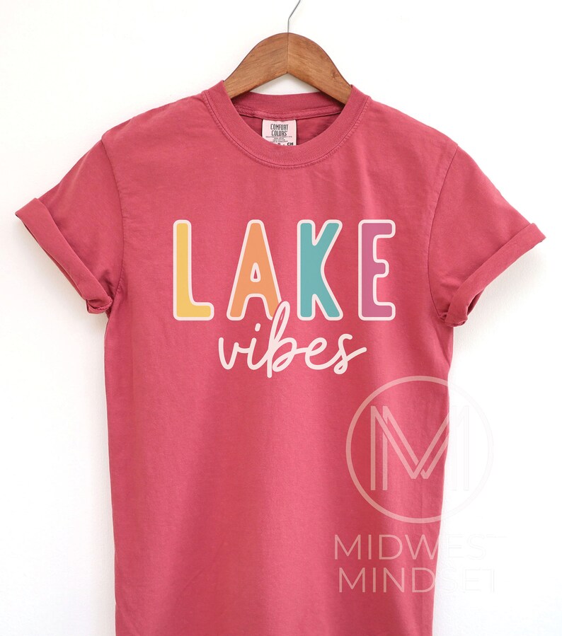 Puede incluir: Una camiseta roja de manga corta con las palabras "LAKE vibes" impresas en la parte delantera en letras coloridas. La camiseta est&aacute; colgada en una percha de madera sobre un fondo blanco. El texto est&aacute; en una fuente l&uacute;dica e informal.