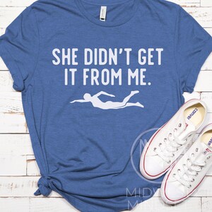 May include: A blue t-shirt with the text "She didn't get it from me." in white. A white silhouette of a swimmer is below the text.