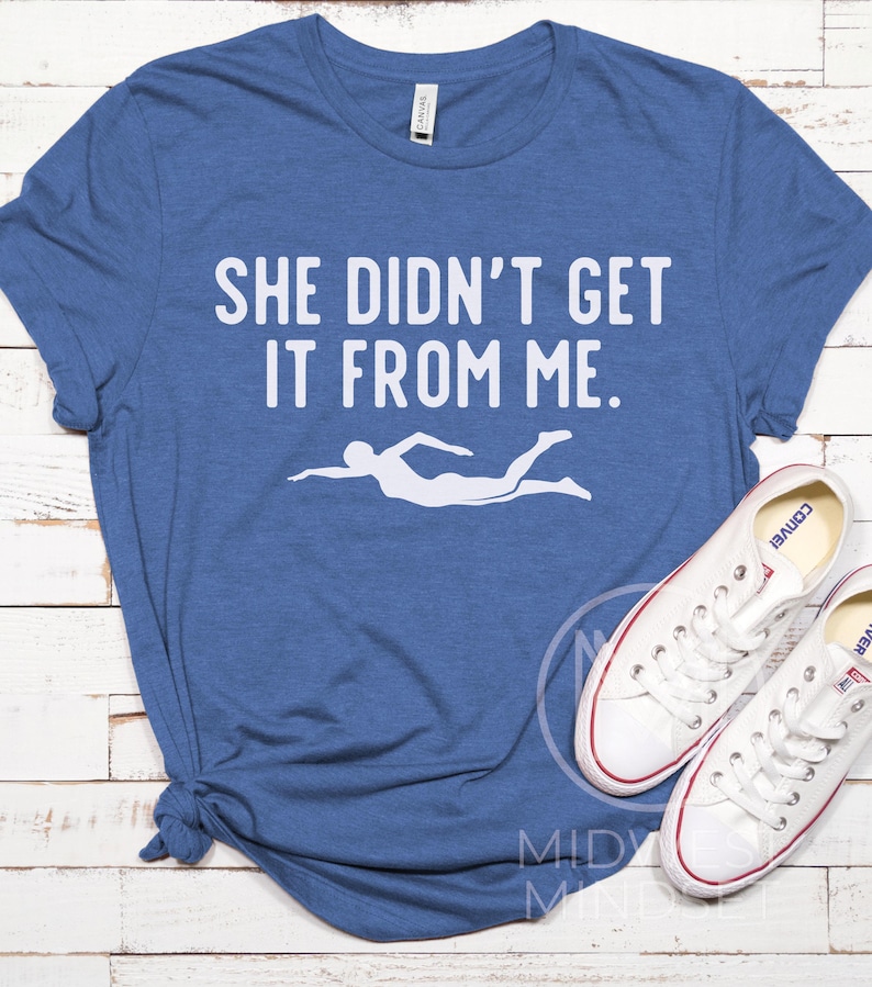 May include: A blue t-shirt with the text "She didn't get it from me." in white. A white silhouette of a swimmer is below the text.