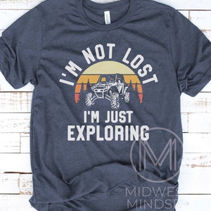 K&ouml;nnte beinhalten: Ein marineblaues T-Shirt mit einer Grafik im Vintage-Stil eines Sonnenuntergangs, B&auml;umen und einem Side-by-Side-Gel&auml;ndewagen. Der Text "Ich bin nicht verloren, ich erkunde nur" ist in Wei&szlig; auf dem Shirt gedruckt.