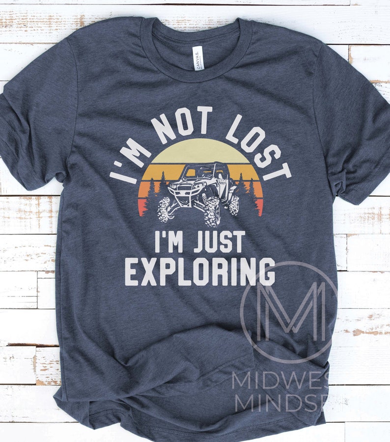 K&ouml;nnte beinhalten: Ein marineblaues T-Shirt mit einer Grafik im Vintage-Stil eines Sonnenuntergangs, B&auml;umen und einem Side-by-Side-Gel&auml;ndewagen. Der Text "Ich bin nicht verloren, ich erkunde nur" ist in Wei&szlig; auf dem Shirt gedruckt.