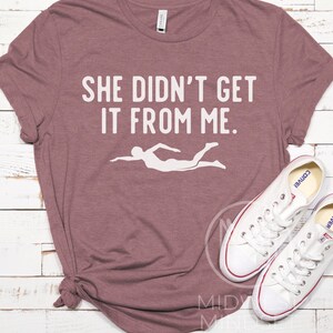 May include: A mauve t-shirt with the text "She didn't get it from me." in white. A white silhouette of a swimmer is below the text.